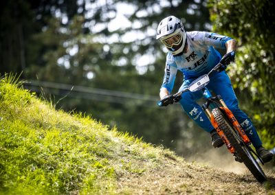 A mountain biker wearing blue and white gear and a helmet rides downhill at speed on a dirt trail surrounded by greenery and trees.