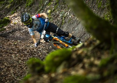 A mountain biker in a blue jersey and black helmet rides quickly around a curve on a forest trail, leaning sharply to the side with leaves and mossy ground in the background.