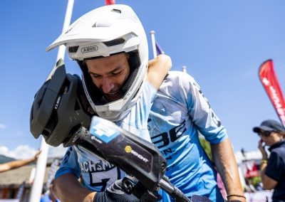 A mountain biker in full gear, including a white helmet, receives a hug from a supporter after a race, with flags and a blue sky in the background.