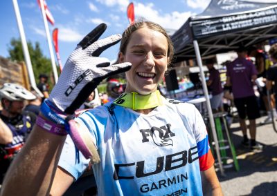 A smiling mountain biker wearing a blue CUBE jersey and gloves holds up three fingers to the camera, with people, bikes, and tents in the background on a sunny day.