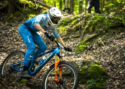 A mountain biker wearing blue gear and a white helmet rides a blue bike down a leafy, forested trail. Sunlight filters through the trees, and moss-covered rocks are visible in the background.