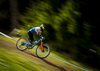 A cyclist in a blue and white jersey rides a mountain bike at high speed on a dirt trail, with motion blur emphasizing the sense of rapid movement and a green, wooded background.