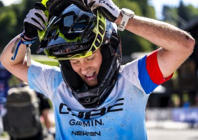 A cyclist wearing a white and blue jersey with sponsor logos lifts off a black helmet, with visible dirt on their arms and shirt, smiling outdoors on a sunny day.