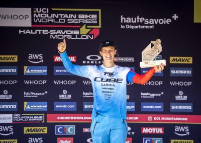 A smiling male mountain biker in a blue racing suit holds a trophy and gives a thumbs up on the podium at the UCI Mountain Bike World Series in Haute-Savoie, Morillon, with event sponsor logos in the background.