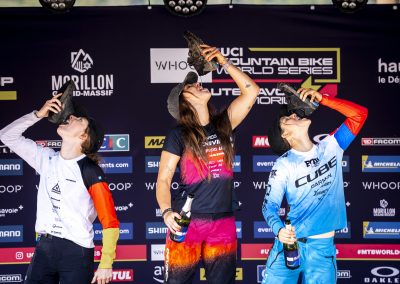 Three mountain bike racers celebrate on a podium by pouring drinks into their shoes and drinking, surrounded by sponsor logos at the UCI Mountain Bike World Series event.