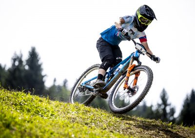 A mountain biker wearing a helmet, goggles, and blue jersey rides downhill on a grassy slope, leaning into the turn with trees and a bright sky in the background.