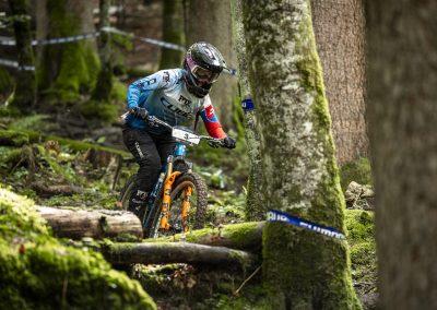 A mountain biker wearing a blue and white jersey and helmet rides down a mossy, forested trail, maneuvering between trees and over uneven terrain during a race.