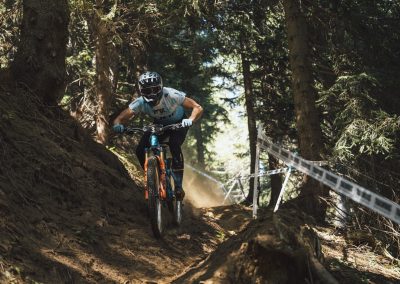A mountain biker wearing a helmet and goggles rides downhill through a steep, narrow dirt trail in a forest, kicking up dust and navigating between trees and a taped course boundary.