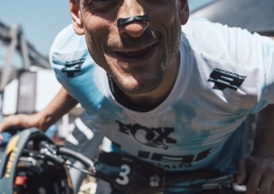 A cyclist in a white jersey with black markings on his nose smiles at the camera while holding a helmet and leaning over a blue bicycle outdoors on a sunny day.