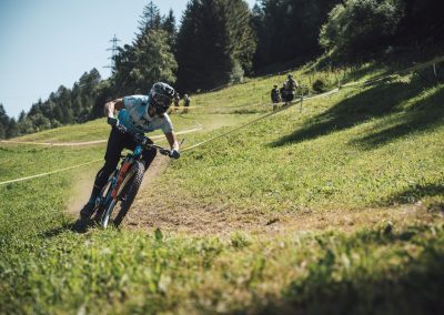 A mountain biker wearing a helmet and sunglasses rides quickly down a grassy trail on a sunny day, surrounded by trees and hills, with a few people in the background.