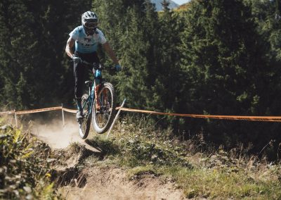 A mountain biker in a blue and white jersey and helmet rides over a dirt trail jump, lifting wheels off the ground, surrounded by trees and orange race tape under a sunny sky.