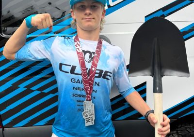 A young person in a blue Shimano cap and Cube cycling jersey stands in front of a branded vehicle, holding a shovel and wearing a medal around their neck while making a proud fist gesture.