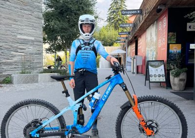 A person wearing a blue and white biking outfit and helmet stands on a paved street holding a blue and orange Cube mountain bike. There are buildings, signs, trees, and a menu board in the background.