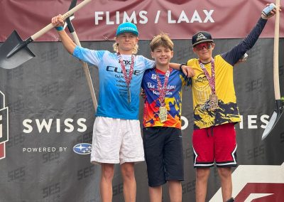 Three boys stand on a podium with medals around their necks, holding shovels. They are smiling and posing in front of a FLIMS / LAAX banner at a Swiss cycling event. A bicycle is visible at the bottom of the image.
