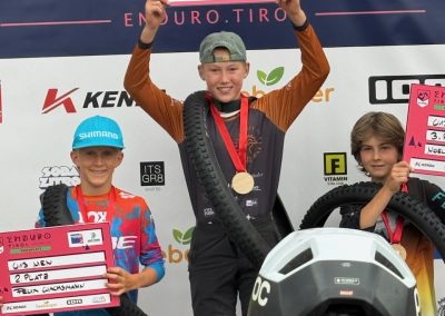 Three boys stand on a podium holding certificates and tires as prizes. The center boy, smiling and wearing a gold medal, stands on the top step. A backdrop displays event sponsors and logos behind them.