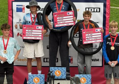 Five teenage boys with medals stand on a podium at a mountain biking event. The top three hold prize boards and bike tires, with a red sponsor backdrop and grass in the background.