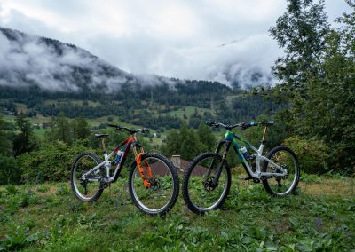 Two mountain bikes are parked on grass, overlooking a lush green valley with scattered trees and houses. Misty clouds hang over forested hills and mountains in the background.
