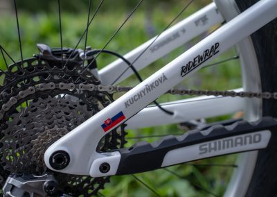 Close-up of a white bicycle frame with gear cassette, chain, and Shimano branding visible. Stickers include the Slovak flag, the name Kuchynková, and the RideWrap logo. Green foliage is blurred in the background.