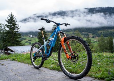 A mountain bike with orange and blue details is parked on a stone path, overlooking misty hills, green fields, trees, and distant mountains partially covered in low clouds.