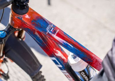 Close-up of a red and blue mountain bike frame with a water bottle mounted on it, showing the Cube brand logo and a Garmin label. The bike is outdoors on a sunny day.