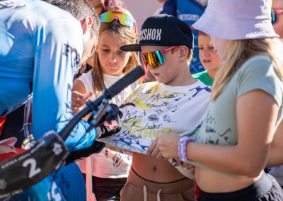 A group of children gather around a cyclist as he signs a T-shirt covered in signatures. One child holds out the shirt, wearing a cap and colorful sunglasses, while others watch closely. A bicycle is partially visible in the foreground.