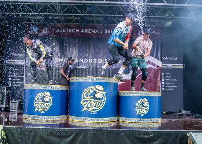 Three people stand on barrels on a stage, with water being sprayed over them. They react with surprise and laughter as water splashes in the air. The background shows event banners and a partially covered stage.