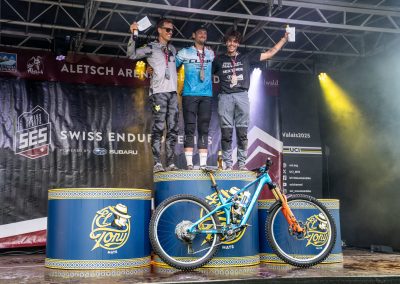 Three male cyclists stand on a winners’ podium with medals and certificates, smiling and celebrating. A bright blue mountain bike is displayed in front. The background shows event banners and stage lights.