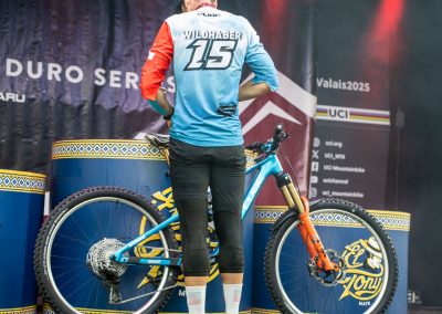 A cyclist in a blue, red, and white jersey with Wildhaber 15 stands on a stage next to a mountain bike, facing away from the camera, surrounded by decorative barrels and event signage.