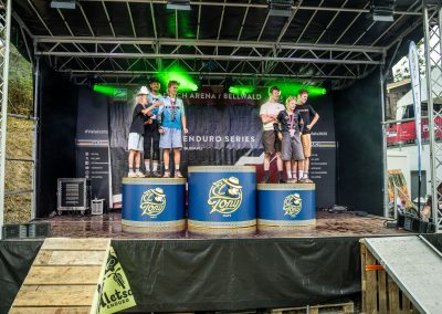 Six cyclists stand on a podium with medals at an award ceremony for the Enduro Series. The podium is under a stage with green lights, and the event banner is visible in the background.