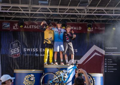 Three mountain bikers stand on a podium celebrating their win at a Swiss Enduro event, with one holding a trophy. Spectators take photos while banners and trees are visible in the background.
