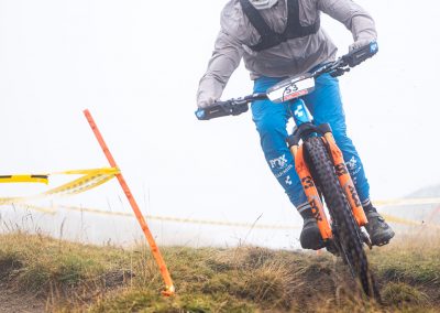 A mountain biker wearing blue pants, a gray jacket, and a helmet rides downhill on a dirt trail, leaning into a turn near orange and yellow trail markers in a grassy, foggy setting.