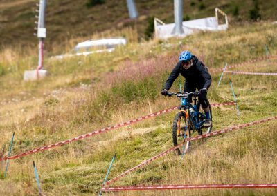 A cyclist wearing a blue helmet and dark clothing rides a mountain bike downhill along a marked trail on grassy terrain during a competition or event.