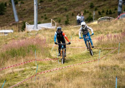 Two mountain bikers ride down a grassy, marked trail during a race, surrounded by wild grass and hills, while a few spectators watch in the background.