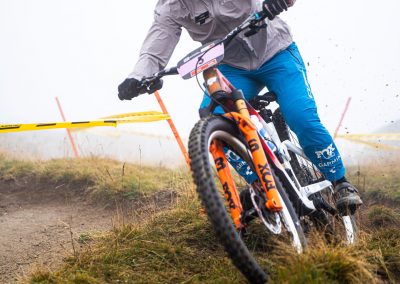A mountain biker in a helmet and goggles rides downhill on a dirt trail, leaning into a turn. The rider wears a gray jacket and blue pants. Grass and some yellow caution tape are visible in the background.