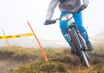 A mountain biker wearing blue pants, a white jacket, and a helmet with goggles rides downhill on a dirt trail marked by yellow caution tape and orange stakes. The background is foggy and grassy.