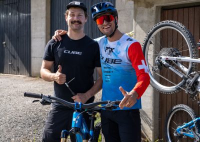 Two men in cycling gear stand outdoors by mountain bikes, smiling and giving thumbs up. One wears a blue and red jersey and helmet, the other a black shirt and cap. A bike is propped up nearby against a concrete building.