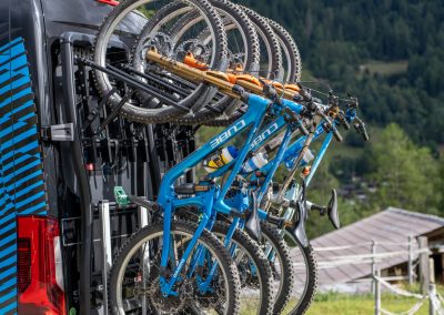 A row of mountain bikes mounted on a vertical bike rack attached to the back of a van, with forested hills and rooftops visible in the background.