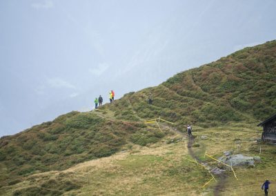 Several hikers walk along a winding trail on a grassy, hilly landscape with low shrubs. Some yellow tape lines parts of the path, and a small wooden shelter sits to the right. The sky is overcast with a misty, hazy background.