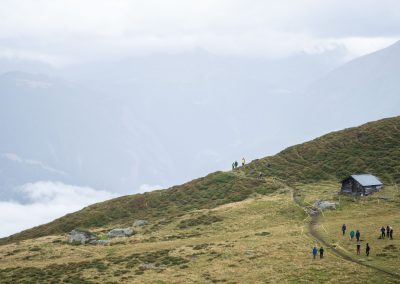 A group of hikers walks along a winding trail on a grassy mountainside with a small wooden cabin nearby and misty mountains in the background.