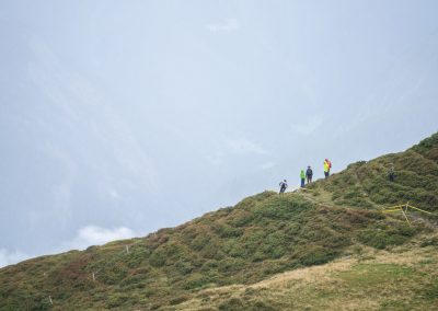 Four hikers walk along a grassy, shrub-covered mountain ridge on a misty day, surrounded by fog and distant, faintly visible hills in the background.
