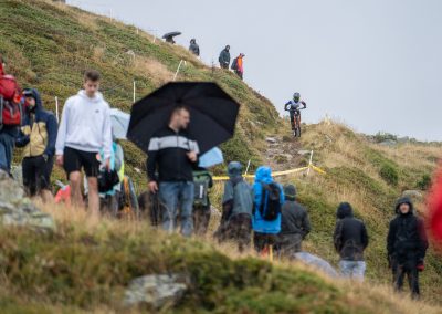 A mountain biker rides down a rocky trail during a race, while spectators in rain jackets and holding umbrellas watch from the grassy hillside on a cloudy, rainy day.