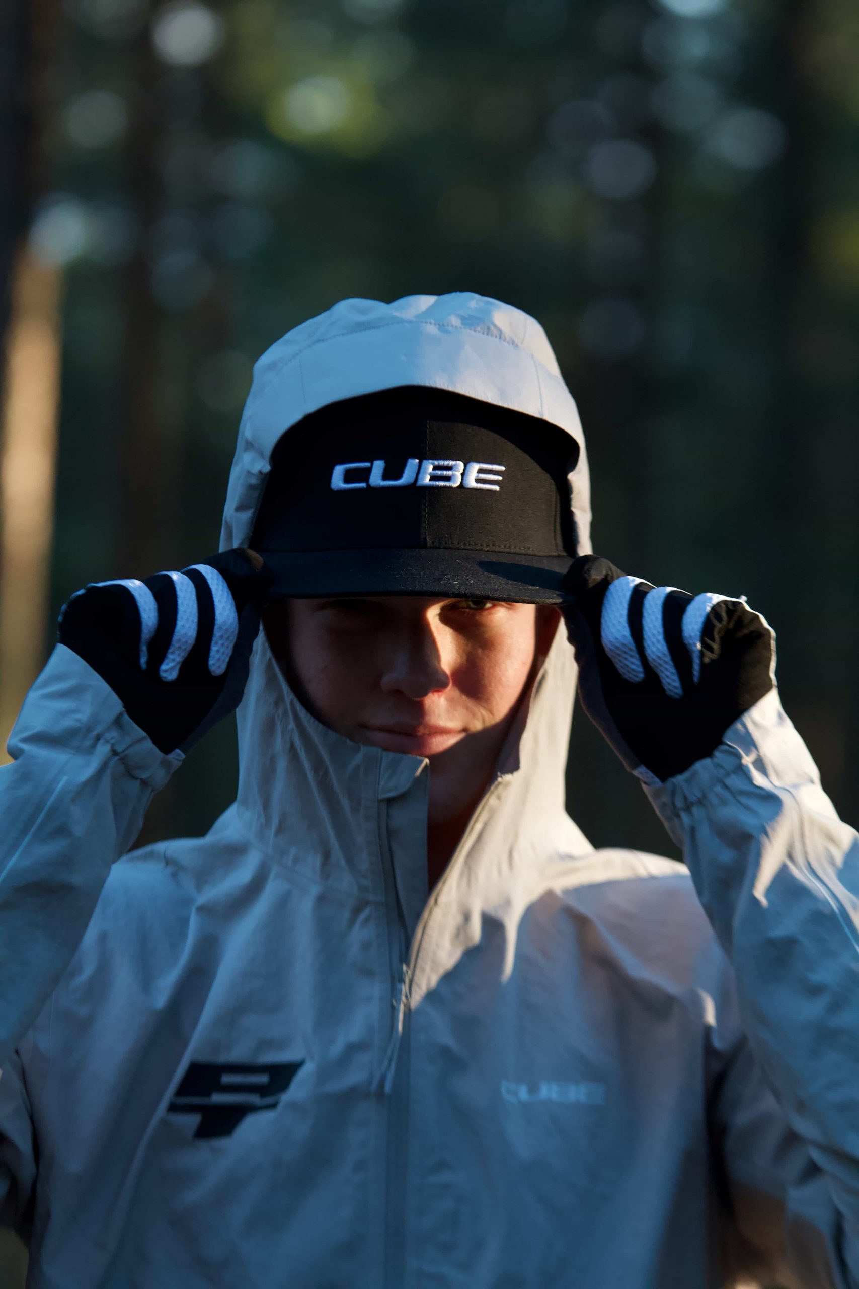 IMG_3523 A person in a light gray jacket with the hood up adjusts a black cap labeled CUBE. They are wearing gloves, and the background is an out-of-focus forest with sunlight filtering through the trees.
