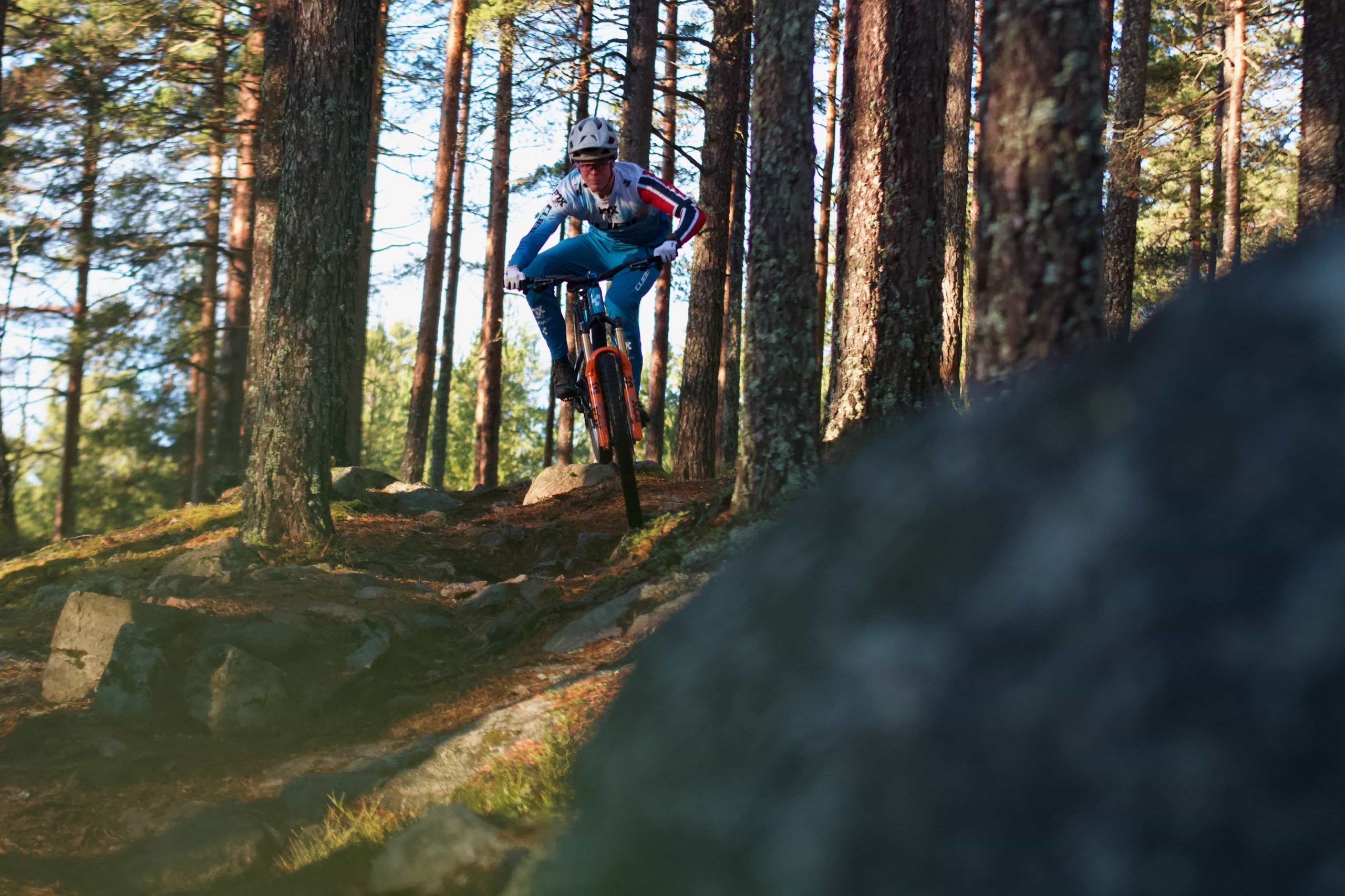 IMG_3556 A person wearing a helmet rides a mountain bike down a rocky trail through a dense forest with tall trees and dappled sunlight. A large, blurry rock is visible in the foreground.