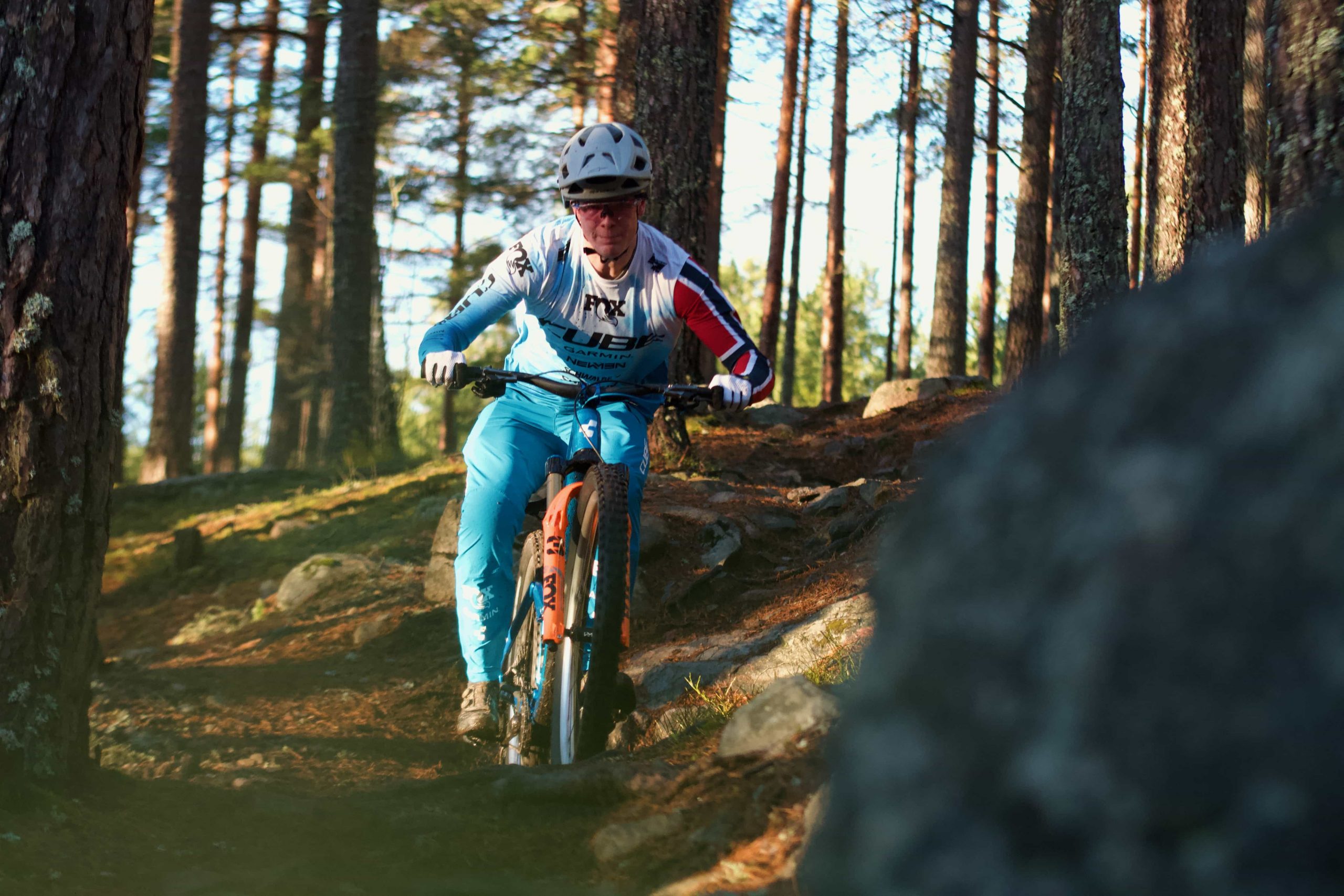 IMG_3564 A cyclist wearing a helmet and blue and white gear rides a mountain bike through a forest trail with trees, rocks, and sunlight filtering through the branches.