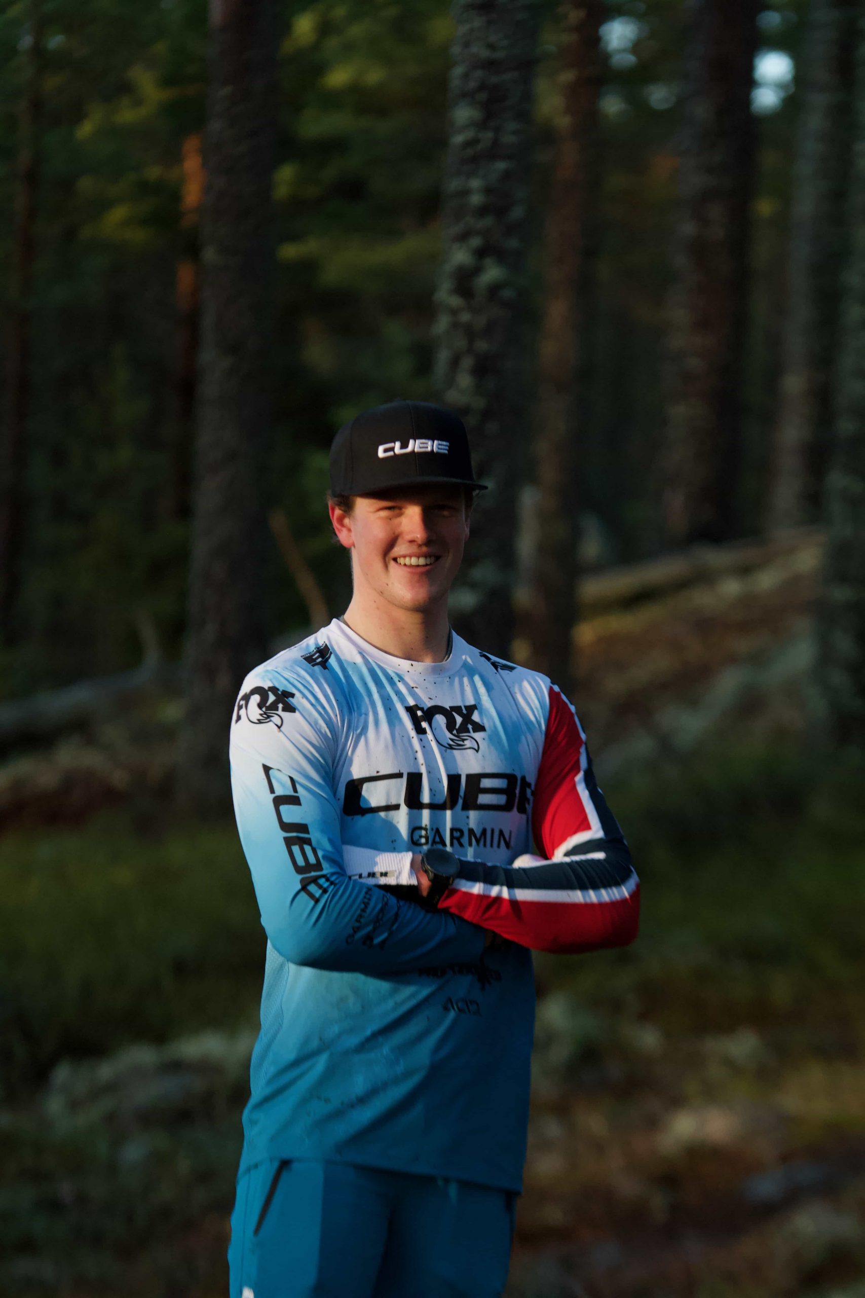 IMG_3604 A man wearing a CUBE cap and a blue cycling jersey with sponsor logos stands smiling with arms crossed in a sunlit forest.
