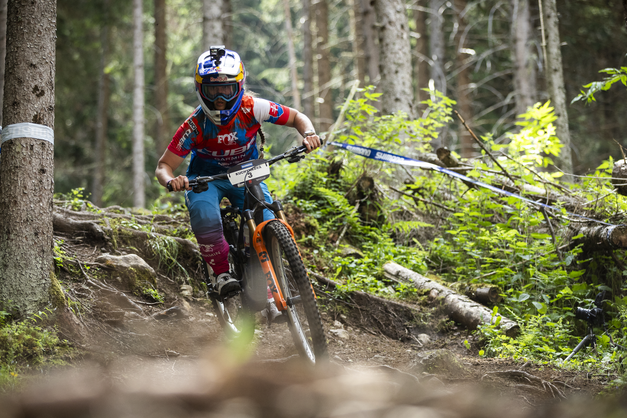 Mountain biker wearing bright gear and a helmet rides downhill on a rocky trail with tall, rugged mountains and a clear blue sky in the background.